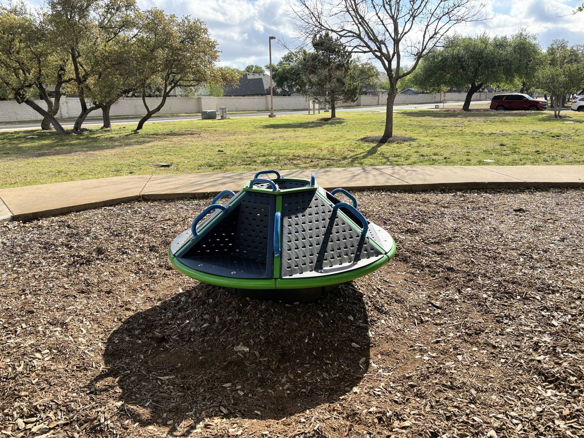 A green and grey circular spinning playground merry-go-round with blue metal handrails sits on a woodchip surface in a park surrounded by trees and a paved path under a bright sky.