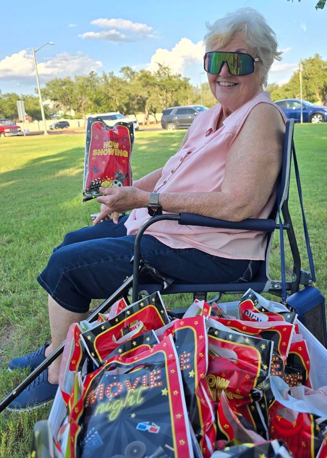 Director Norrell smiles while seated in a folding chair at an outdoor movie event. She holds a themed party favor bag, with a bin full of additional "Movie Night" bags resting on the grass in front of her.