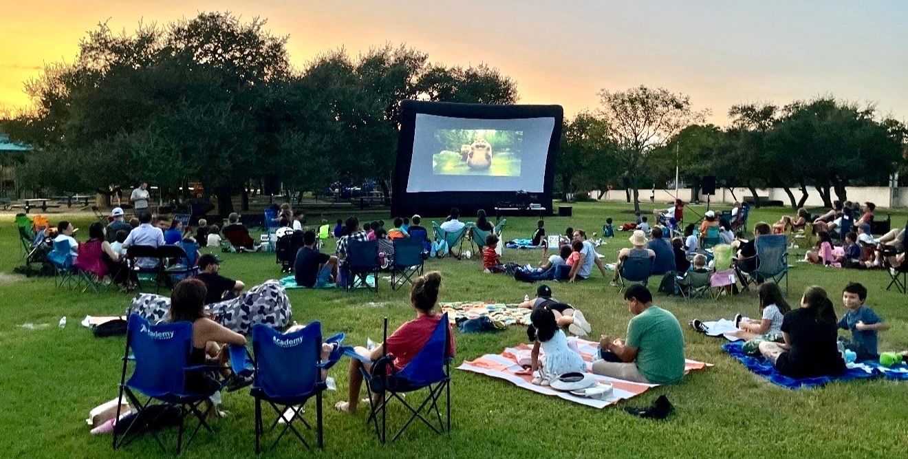 People sitting on blankets and folding chairs in a grassy park, watching an outdoor movie projected onto a large screen at dusk