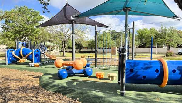 Modern playground with blue and orange tunnels, climbing ropes, and shade sails over a green rubberized surface. A wooden gazebo and lush green trees are visible in the background under a bright sky.